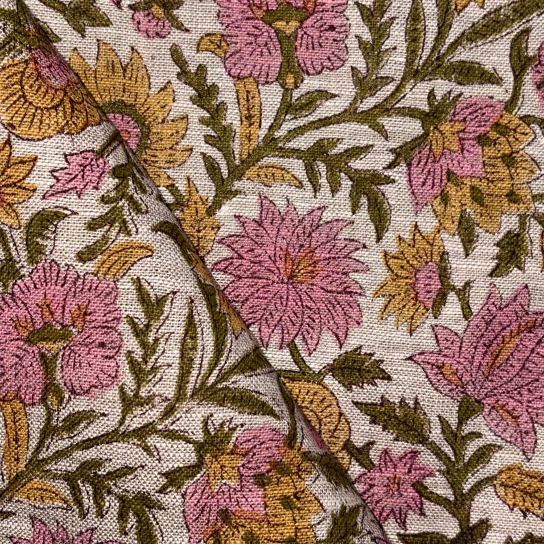 Op de afbeelding: Close-up van een bloemenstof met een herhalend patroon van roze en gele bloemen, groene bladeren en stelen op een cr&egrave;mekleurige achtergrond. De stof is gevouwen, waardoor de continu&iuml;teit van het patroon zichtbaar is. Het ontwerp heeft een vintage of bohemien esthetiek.