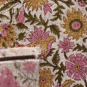 Op de afbeelding: Close-up van bloemenstof met een herhalend patroon van roze en gele bloemen en groene bladeren op een cr&egrave;mekleurige achtergrond. De stof heeft een rafelige rand en een gestreepte rand.