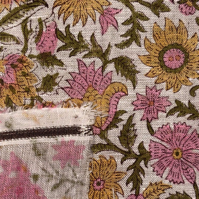 Op de afbeelding: Close-up van bloemenstof met een herhalend patroon van roze en gele bloemen en groene bladeren op een cr&egrave;mekleurige achtergrond. De stof heeft een rafelige rand en een gestreepte rand.
