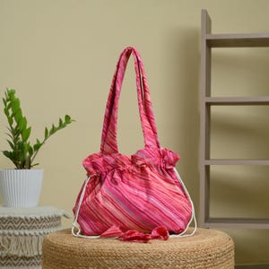 May include: A pink tote bag featuring a diagonal striped pattern in various shades of pink, red, and white. The bag has two shoulder straps, a gathered top opening, and white drawstring cords that hang down the front with small fabric knots.