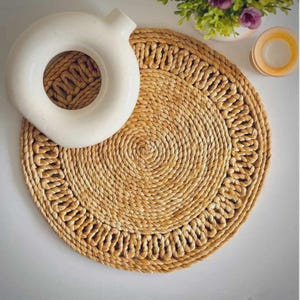 Puede incluir: Un salvamanteles redondo tejido con un diseño en espiral y un borde decorativo. Un jarrón de cerámica blanco en forma de anillo descansa sobre el salvamanteles. El salvamanteles está hecho de fibras naturales y es de color marrón claro.
