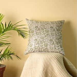 May include: Square cushion featuring a floral design in green and white. The cushion is placed on a cream-coloured blanket. A potted plant with green leaves is in the background.