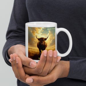 Könnte beinhalten: Weiße Keramiktasse mit einer Stier-Illustration vor goldenem Himmel. Auf der Tasse steht "April 20 - May 20" und der Buchstabe "T" auf der Vorderseite. Die Tasse wird von einer Person gehalten.