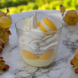 Puede incluir: Una vela de cristal transparente con diseño de postre. La vela presenta capas de cera blanca, cera amarilla y un macaron amarillo. El texto "HANDMADE CANDLES" y "BY Vivien" está impreso en la vela.