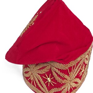 May include: A red velvet hat with intricate gold embroidery. The hat features a square base with detailed floral patterns and a pointed top. The gold embroidery is made of small beads and sequins. The hat is likely a ceremonial or decorative piece.