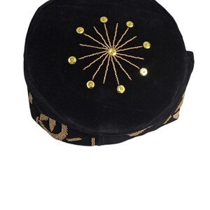 May include: Black velvet cap with gold-colored embellishments. The cap features a sunburst design made of gold beads and sequins on the crown. The brim is also decorated with gold embroidery. This headwear is suitable for special occasions.