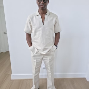 May include: A cream-colored linen outfit consisting of a short-sleeved collared shirt with black trim and matching pants. The outfit is worn with black shoes and sunglasses. The shirt has a V-neck design.