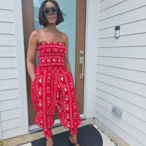 May include: A strapless jumpsuit in a vibrant red fabric featuring a repeating white geometric pattern. The garment includes a smocked bodice, a cinched waist, and loose-fitting trousers with tapered cuffs at the ankles and side pockets.