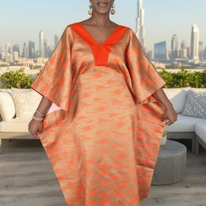 May include: An orange and tan caftan dress with a V-neckline and wide sleeves. The dress features an all-over pattern of orange shapes. The model is wearing a white head wrap and gold earrings. The background shows a cityscape.