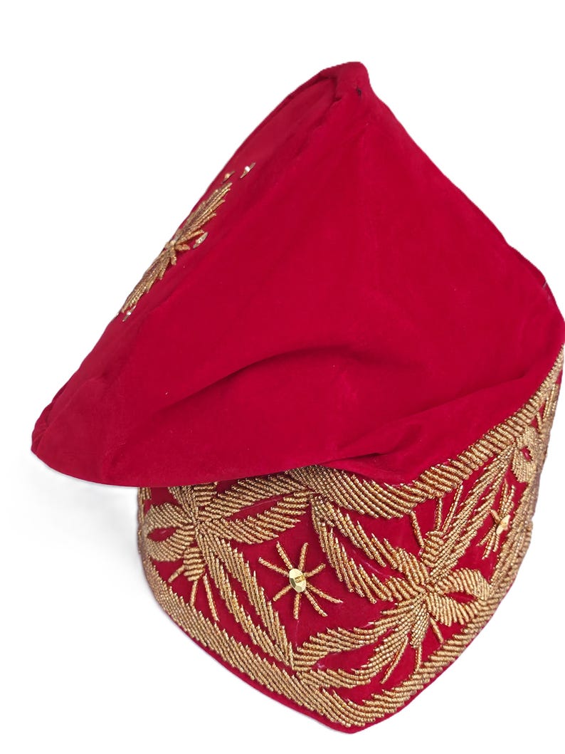 May include: A red velvet hat with intricate gold embroidery. The hat features a square base with detailed floral patterns and a pointed top. The gold embroidery is made of small beads and sequins. The hat is likely a ceremonial or decorative piece.