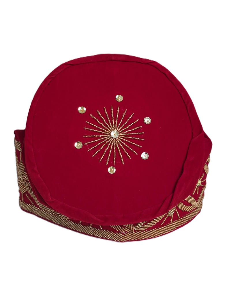 May include: A red velvet hat with gold beaded trim and a gold embroidered starburst design on top. The hat is a traditional style, likely for a special occasion or cultural event.