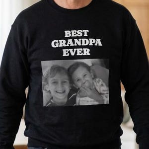 Peut inclure: Sweat-shirt noir avec l'inscription blanche "BEST GRANDPA EVER". En dessous, une photo en noir et blanc de deux enfants souriants. Le sweat-shirt est de style col rond.