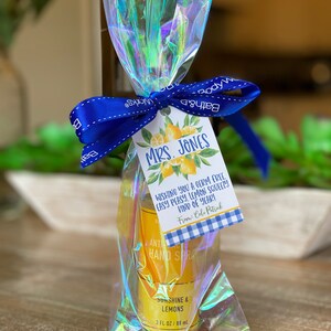 May include: A clear plastic bag with a blue ribbon tied around it. Inside the bag is a small bottle of hand sanitizer with a yellow label that says "Sunshine & Lemons" and "3 fl oz / 88 ml". A small tag with a lemon design and the text "Mrs. Jones Wishing you a germ free, easy peasy, lemon squeezy kind of year! From: Colo Patrick" is attached to the ribbon.