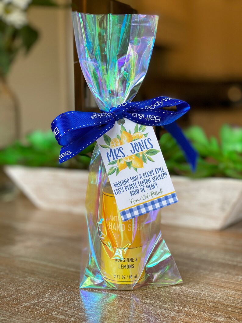 May include: A clear plastic bag with a blue ribbon tied around it. Inside the bag is a small bottle of hand sanitizer with a yellow label that says "Sunshine & Lemons" and "3 fl oz / 88 ml". A small tag with a lemon design and the text "Mrs. Jones Wishing you a germ free, easy peasy, lemon squeezy kind of year! From: Colo Patrick" is attached to the ribbon.