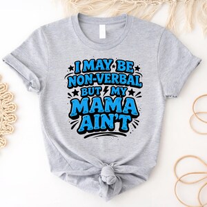 Puede incluir: Camiseta gris claro con el texto "I MAY BE NON-VERBAL BUT MY MAMA AIN'T" en azul y negro. El texto está estilizado con estrellas y rayos. La camiseta está anudada en la parte inferior.