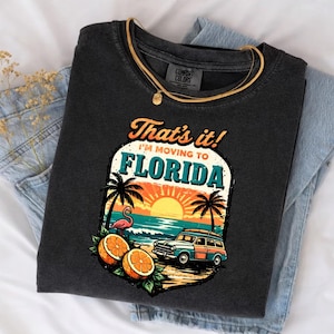 Może przedstawiać: Ciemnoszara koszulka z napisem "That's it! I'm moving to Florida" oraz grafiką zachodu słońca, palm, flaminga, pomarańczy i zabytkowego samochodu. Złoty naszyjnik jest zarzucony na koszulkę.