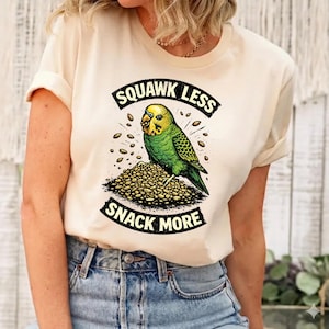 May include: Cream-colored t-shirt featuring a green and yellow parakeet illustration perched on a pile of seeds. The text "SQUAWK LESS" and "SNACK MORE" are printed above and below the bird in a black, curved font. The shirt is worn by a person.