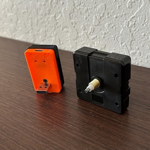 Puede incluir: Dos componentes de reloj se muestran sobre una superficie de madera. Uno es un movimiento de reloj rectangular negro con un eje dorado. El otro es un dispositivo rectangular naranja y negro con un conector USB.