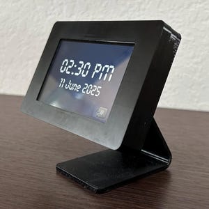 May include: A black digital clock displaying "02:30 PM" and "11 June 2025" on its screen. The clock has a rectangular frame and a stand, and is set on a wooden surface.