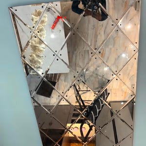 Op de afbeelding: Een grote rechthoekige wandspiegel met een decoratief ruitvormig rasterpatroon over het oppervlak. De spiegel is samengesteld uit meerdere individuele glaspanelen die samen een reflecterend geometrisch ontwerp vormen met kleine donkere accenten.