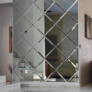 Op de afbeelding: Een grote wandspiegel bestaande uit een raster van ruitvormige glaspanelen met afgeschuinde randen. Het reflecterende oppervlak beslaat een aanzienlijk deel van de muur en creëert een herhalend geometrisch patroon dat de binnenruimte weerspiegelt.