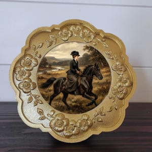 May include: A gold-colored, floral-edged decorative plate featuring a vintage painting of a person riding a black horse through a landscape. The rider is dressed in a dark outfit and hat. The plate has a scalloped edge.