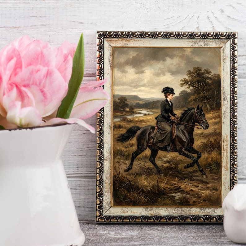 May include: An antique framed print depicts a woman riding a black horse through a field. The artwork features a woman in a dark riding outfit and hat. The frame is ornate with a cream and gold finish. Pink tulips in a white pitcher are in the foreground.
