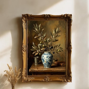 May include: A framed oil painting depicting a blue and white ceramic vase filled with olive branches on a wooden table. The scene includes a metal cup and a leather-bound book, all enclosed in an ornate, antique-style gold-colored frame.