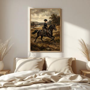 May include: A framed vintage painting depicts a woman riding a black horse across a field. The rider wears a dark dress and hat. The background shows a landscape with a river and trees under a cloudy sky. The artwork is displayed above a bed.