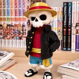 Puede incluir: Figura de un personaje con rostro de calavera que lleva un sombrero de paja, una chaqueta negra, una camisa roja, pantalones cortos azules y sandalias. La figura tiene una faja dorada y está de pie frente a una estantería con mangas y libros abiertos.