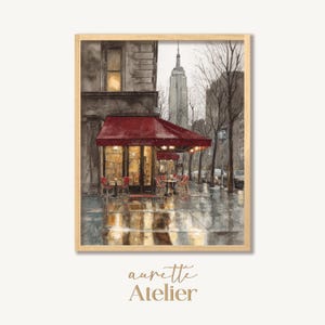 May include: Watercolor painting of a rainy day in New York City, featuring a cafe with a red awning and tables. The Empire State Building is visible in the background. The artwork is framed in a light wood frame, with the text "aurette Atelier" at the bottom.