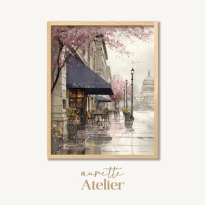 May include: Watercolor print of a cafe scene with a blue awning that reads "Cafe Sire". The artwork depicts a rainy day with pink flowering trees, street lamps, and a building in the background. The print is framed in a light wood frame.