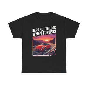 May include: Black t-shirt featuring a graphic of a red convertible on a winding road at sunset. The text "HARD NOT TO LOOK WHEN TOPLESS" is above the car. The design is centered on the shirt.