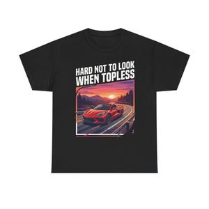 Puede incluir: Camiseta negra con un gráfico de un coche deportivo rojo en una carretera sinuosa al atardecer. El texto "HARD NOT TO LOOK WHEN TOPLESS" está impreso encima de la imagen.