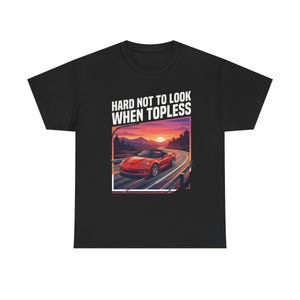 Puede incluir: Camiseta negra con un gráfico de un coche deportivo descapotable rojo en una carretera sinuosa al atardecer. El texto "HARD NOT TO LOOK WHEN TOPLESS" está encima.