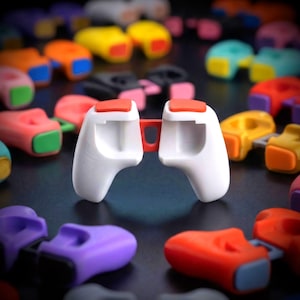May include: A white plastic controller grip accessory featuring two ergonomic handles connected by a central bridge, with bright orange buttons positioned on the top of each handle, surrounded by a collection of similar colourful grips.