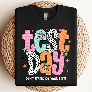 May include: A black crewneck sweatshirt featuring the text "test day" in large, colorful, patterned letters including pink chevron, teal checkerboard, black and white cow print, and orange stripes, with "DON'T STRESS! DO YOUR BEST!" below.