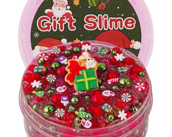 May include: A clear plastic pot filled with red slime, decorated with green and red beads, and festive charms. The lid features a Santa Claus illustration and the words "Gift Slime".
