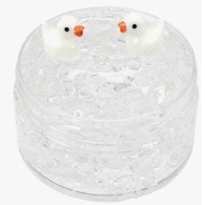Puede incluir: Un recipiente redondo y transparente lleno de pequeños trozos transparentes de forma irregular. Dos pequeñas figuras de pato blanco con picos naranjas y ojos negros se asientan sobre el contenido. El recipiente tiene una tapa transparente y redondeada.