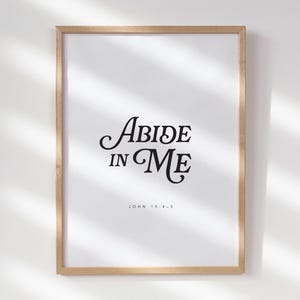 May include: A framed print with the words "Abide in Me" in elegant black script. The text is centered on a white background, with the reference "John 15:4-5" below. The frame is a light brown color.