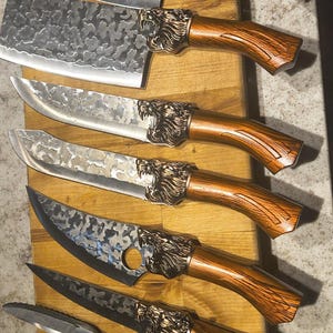 May include: An assortment of handcrafted knives with wooden handles and lion head accents. The blades have a textured finish. Includes a cleaver, chef's knives, and shears, all arranged on a wooden cutting board.