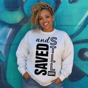 Puede incluir: Una sudadera blanca con las palabras "SAVED" y "STILL DOPE" en letras negras. La sudadera la lleva una persona con cabello rubio rizado y aros dorados, de pie frente a una pared pintada de color turquesa y azul.