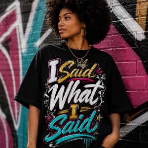 May include: A black short-sleeved t-shirt featuring a graphic print with the text I Said What I Said in stylised white, gold, and teal lettering. The design includes a small gold crown icon above the text and decorative star accents.