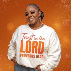 May include: Cream-colored sweatshirt with the text "Trust in the LORD" in red and orange, with "PROVERBS 16:20" below. The sweatshirt is worn by a person with gold hoop earrings and a gold necklace, smiling against an orange background.