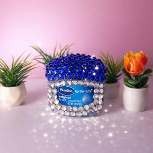 Könnte beinhalten: Ein mit blauen und klaren Strasssteinen verziertes Lippenpflege-Glas. Das Glas hat ein blaues Etikett mit dem Text "Vaseline, lip therapy" und "original for soft, smooth lips". Das Glas ist von kleinen Topfpflanzen und einer dekorativen Blume umgeben.