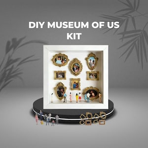 May include: A white shadow box with gold-framed miniature photos, labeled "DIY MUSEUM OF US KIT." Small figurines stand in front of the box. Additional gold frames are scattered below. The box sits on a black and white platform.