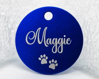May include: A round, royal blue pet tag with the name "Maggie" in white script. Two white paw prints are below the name. The tag has a small hole at the top for attachment.