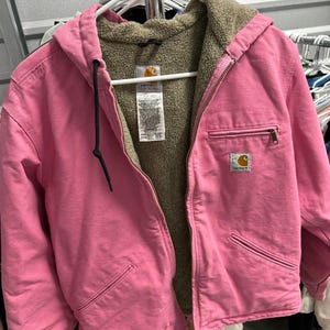 May include: A pink Carhartt hooded jacket with a brown sherpa lining. The jacket has a full zipper, front pockets, and the Carhartt logo patch. The jacket is hanging on a white hanger.