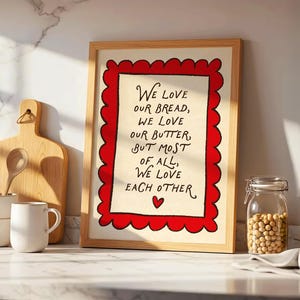 Puede incluir: Una lámina decorativa enmarcada con fondo color crema y un borde rojo festoneado. El texto dice: We love our bread, we love our butter, but most of all, we love each other, con un pequeño icono de corazón rojo centrado en la parte inferior.