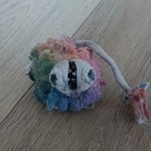 May include: A handmade plush toy with a rainbow-coloured body and two large, grey eyes. A long, grey appendage extends from the body, ending in a frayed, pink and red section. The toy is set against a wooden background.